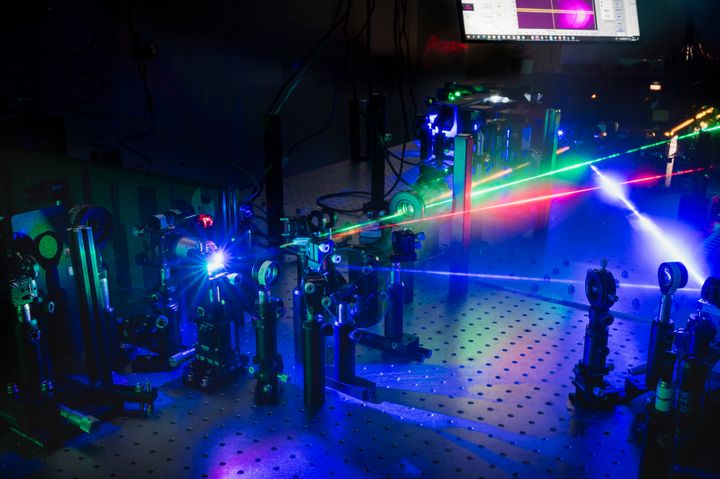 Pimeässä laserlaboratoriossa kuvattu laserlaitteisto, jossa näkyy sinisiä, punaisia ja vihreitä lasersäteitä kuvan poikki. Kuvan laitteisto näkyy sinisessä lasersäteiden synnyttämässä hohteessa, ja tausta on pimeä.