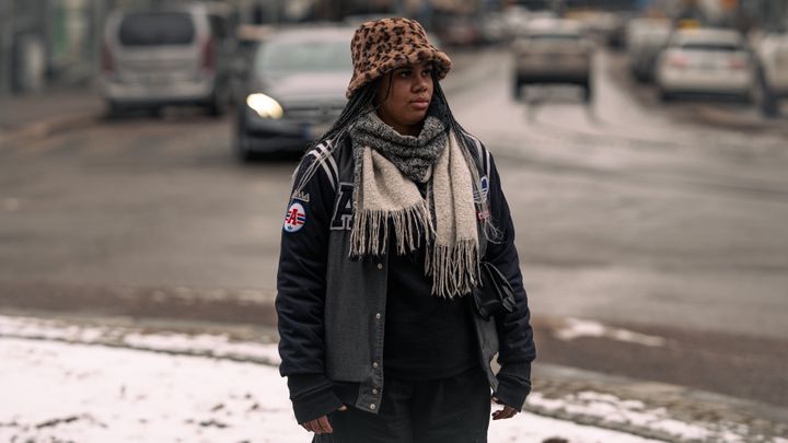 Kuvassa kokemusasiantuntija Amina seisoo talvisella kadulla kaupungissa. Hän katsoo sivulle vakavana ja on pukeutunut lämpimiin talvivaatteisiin.