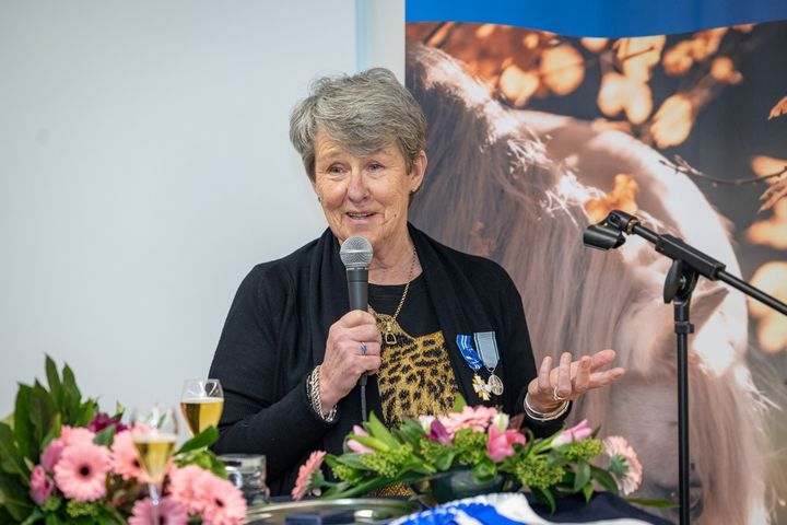 Kyra Kyrklund pitää puhetta mikrofoniin, rinnassaan ansiomerkki, taustalla hevosen kuva ja edessä kukkakoristeita.