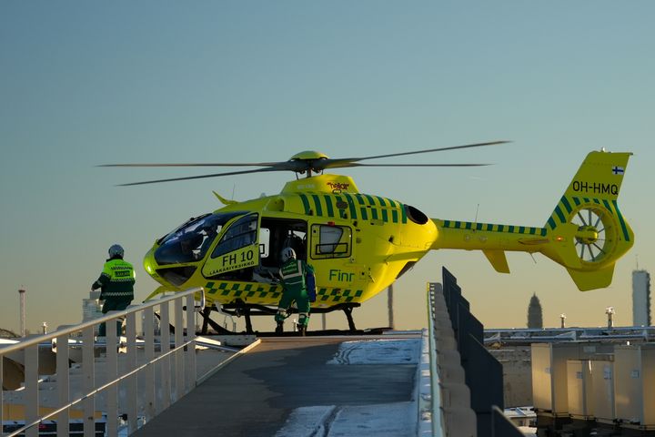 Keltainen lääkärihelikopteri laskeutuneena sairaalan kattokentälle. Kaksi miehistön jäsentä lähestyy helikopteria.