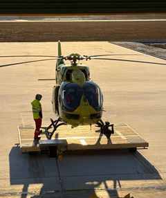 Keltainen lääkärihelikopteri lavetilla Kouvolan tukikohdan edustalla. Vieressä seisoo suojahaalareihin pukeutunut HEMS-ensihoitaja