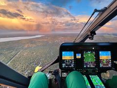 Helikopterin ohjaamosta näkyy tuulilasin läpi laaja metsäalue, järvi sekä auringonlaskun värjäämä taivaanranta. Kuvassa näkyy myös helikopterin mittaristoa ja ohjauslaitteita sekä lentävän miehistön jäsenen jalat haalareissa.