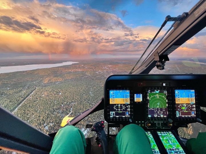 Helikopterin ohjaamosta näkyy tuulilasin läpi laaja metsäalue, järvi sekä auringonlaskun värjäämä taivaanranta. Kuvassa näkyy myös helikopterin mittaristoa ja ohjauslaitteita sekä lentävän miehistön jäsenen jalat haalareissa.