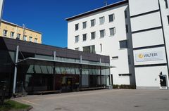 Oikealla viisikerroksinen valkoinen rapattu kivirakennus, johon yhdistyy matalampi tumma lisäosa. Pääoven yläpuolella ja valkoisen rakennuksen seinällä keltainen logopallo ja suurella teksti Valteri.