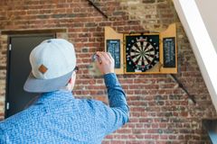 Siniseen paitaan ja harmaaseen taaksepäin käännettyyn lippalakkiin pukeutunut nuori henkilö heittää tikan darts-tauluun.