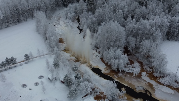 Ilmakuva: Kiurujoki talvimaisemassa, hyytöpadon räjäytys hetki pysäytettynä, vesisuihkut kohoavat taivaalle.