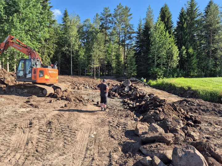 Kaivinkone työskentelee maastossa suuren kivikasan vieressä, taustalla on metsää ja puita.