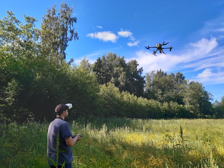Henkilö ohjaa ilmassa lentävää dronea vehreällä niittyalueella. Taustalla näkyy lehtipuita ja sininen, osittain pilvinen taivas.