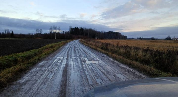 Sorainen tie peltomaiseman keskellä, märkä ja mutainen pinta, horisontissa metsää ja pilvinen taivas iltahämärässä.