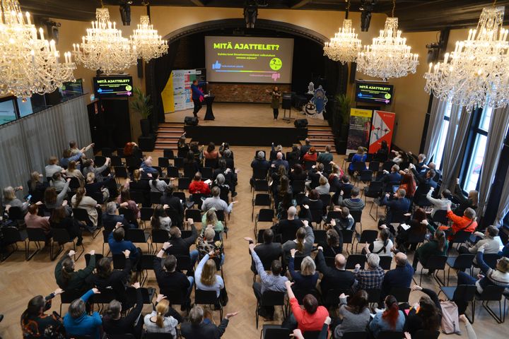 Salillinen osallistujia täydessä hotelli Puijonsarven salissa äänestämässä peukulla.