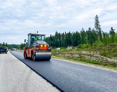 Tietä päällystetään jyrällä. Taustalla on metsää ja muutama auto liikenteessä.