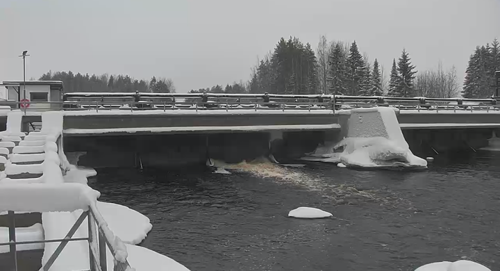 Lumipeitteinen pato, jonka alapuolella vesi virtaa talvisessa maisemassa. Taustalla näkyy lumisia puita.