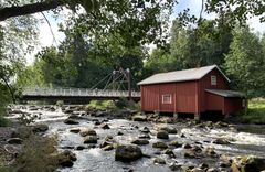 Punainen mökki joen rannalla metsäisessä ympäristössä, taustalla silta.