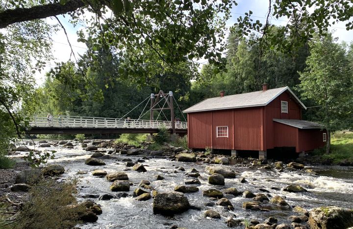 Punainen mökki joen rannalla metsäisessä ympäristössä, taustalla silta.