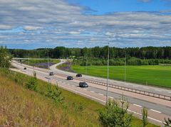 Moottoritie, jolla ajaa useita autoja, ympärillä vihreää metsää ja peltoa. Taustalla sininen taivas ja pilviä.
