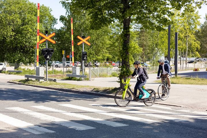 Kaksi pyöräilijää kypärät päässä ylittämässä suojatietä rautatien tasoristeyksessä aurinkoisena päivänä.