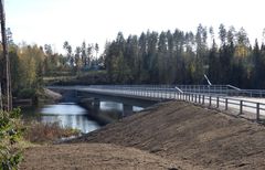 Silta ylittää metsäisen joen, vieressä sorainen tie ja kaiteet.