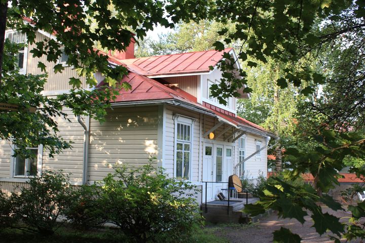 Vaaleankeltainen puutalo punaisella peltikatolla, ympärillä vihreää puustoa ja pensaita.
