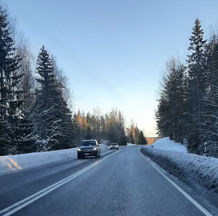 Autoja ajamassa lumisella maantiellä, jonka molemmilla puolilla on kuusimetsää ja kirkas sininen taivas.