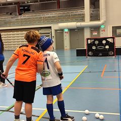 Kaksi nuorta poikaa selin kameraan sisäpelivarusteissa salibandykentällä.