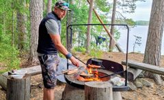 Iäkäs mies paistaa makkaraa retkipaikan nuotiolla metsässä, taustalla järvi.