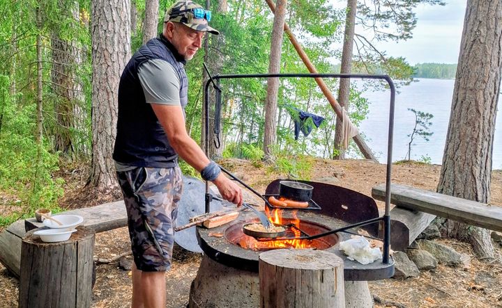 Iäkäs mies paistaa makkaraa retkipaikan nuotiolla metsässä, taustalla järvi.