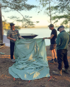 Kolme miestä pystyttää teltaa kesäiltana järven rannalla.