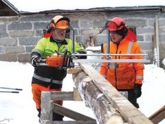 Kaksi miestä työsuojavaatteissa sahaa lankkua ulkona, maassa lunta.