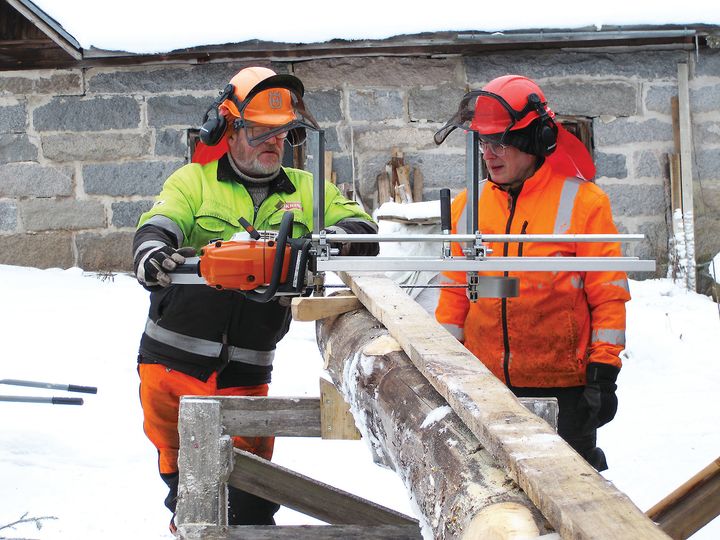 Kaksi miestä työsuojavaatteissa sahaa lankkua ulkona, maassa lunta.