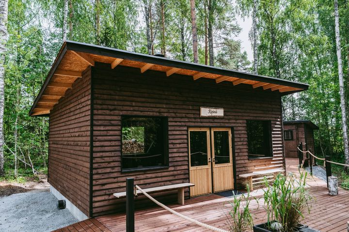 Koivujen katveessa sijaitseva tummasta laudasta tehty ulkorakennus, jossa puinen terassi