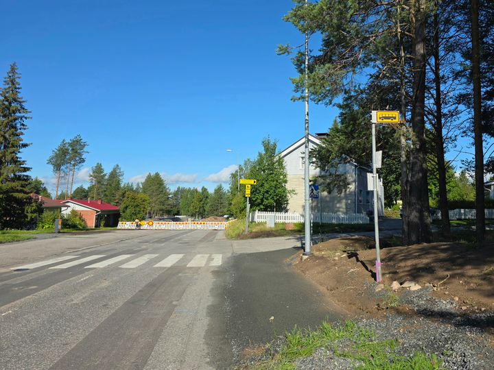Bussipysäkki tien vasemmassa reunassa, taustalla taloja ja työmaa-aidoin suljettu tie.