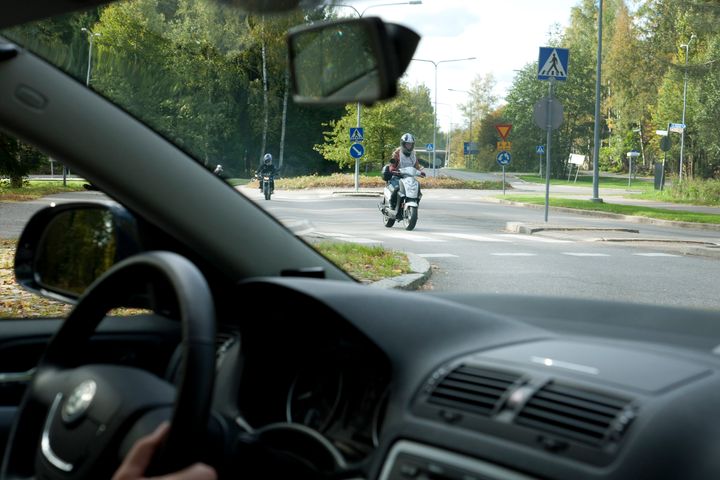 Etualalla auton tuulilasi, jonka läpi näkymä liikenneympyrään, jossa ajaa mopoilijoita.