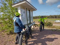 Pyöräilijöitä opastekyltin edustalla aurinkoisessa säässä.