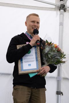 Kai Alajoki puhuu mikrofoniin sylissään kukkakimppu ja kunniakirja.