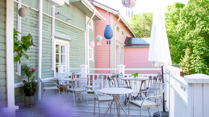 En solig terrass mellan pastellfärgade trähus med små bord, stolar, ett vitt parasoll och hängande lyktor.