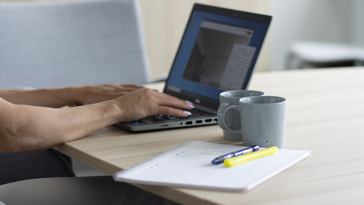 Nainen tietokoneella. Kaksi kahvikuppia taustalla ja paperia sekä kynä.