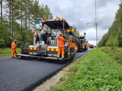 Tienpäällystyskoneita ja niitä käyttävät työntekijät oransseissa turva-asuissa työskentelevät maantiellä vehreän metsän reunalla.