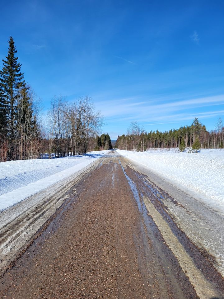 Kelirikkoinen soratie kulkee lumisen maiseman halki, taustalla metsää ja sinistä taivasta.
