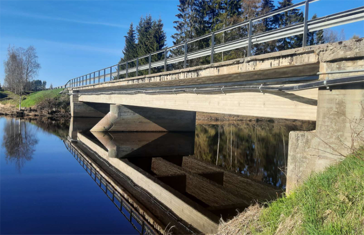Sillan rakenne ja kaiderakenteet, sininen taivas ja ympäröivä luonto heijastuvat veden pinnasta.