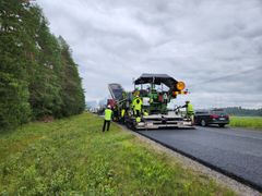 Tietyömaa, jossa useita työntekijöitä asvaltoimassa maantietä metsän vieressä.