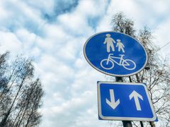 Sininen liikennemerkki, jossa on jalankulkijan ja pyöräilijän symbolit. Merkin taustalla lehtipuita ja pilvinen taivas.