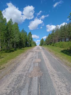 Huonokuntoinen päällystetty tie, ympärillä matsää ja taustalla kesäinen sininen taivas, jossa pilviä.
