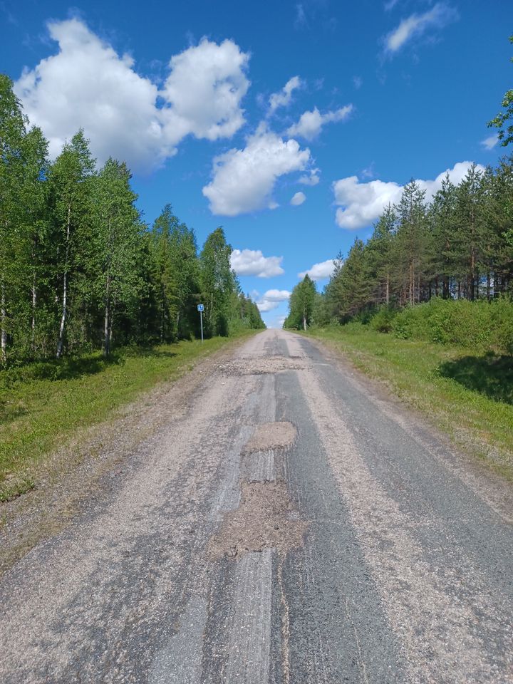 Huonokuntoinen päällystetty tie, ympärillä matsää ja taustalla kesäinen sininen taivas, jossa pilviä.