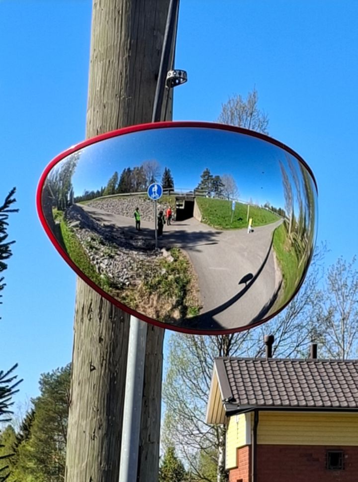 Pyöräilijöitä ja jalankulkijoita näkyy liikennepeilistä aurinkoisella tiellä.