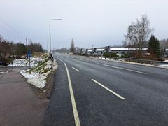 En lång, asfalterad väg med räcken på sidan. Det syns snö längs vägkanten och några träd i bakgrunden. Himlen är grå.