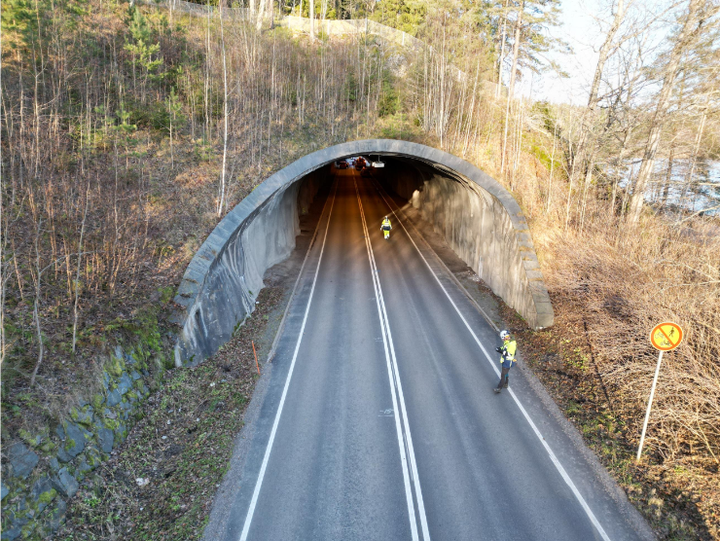 Maantie kulkee metsäisen tunnelin läpi, jossa kaksi ihmistä liikkuu valoisassa päiväsaikaan.
