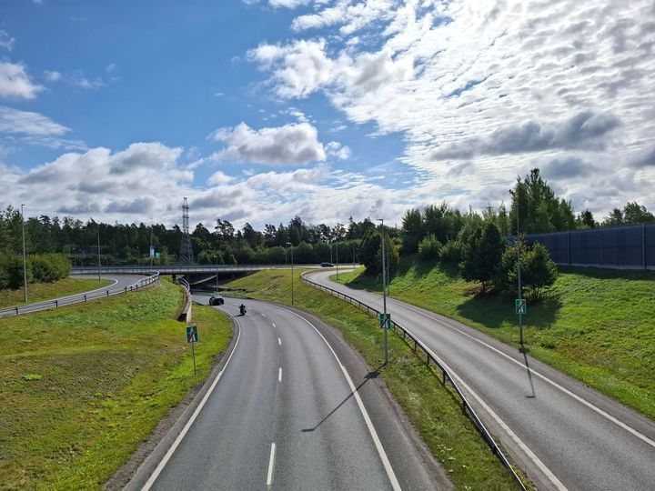 Kuvassa Valtatien 1 ja Kehä II:n eritasoliittymän (Leppäsolmu) ramppi, jossa näkyy autoja ajamassa. Taustalla vehreää maisemaa ja pilvinen taivas.