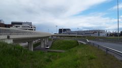 Alt text: Laaja näkymä Hannukaisen risteyssillasta Vantaalla. Etualalla on silta ja sen alapuolella kulkeva tie, taustalla rakennuksia ja pilvinen taivas.