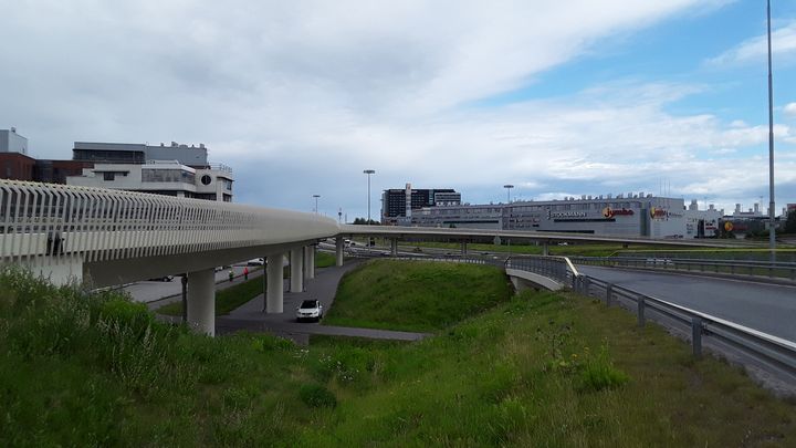 Alt text: Laaja näkymä Hannukaisen risteyssillasta Vantaalla. Etualalla on silta ja sen alapuolella kulkeva tie, taustalla rakennuksia ja pilvinen taivas.