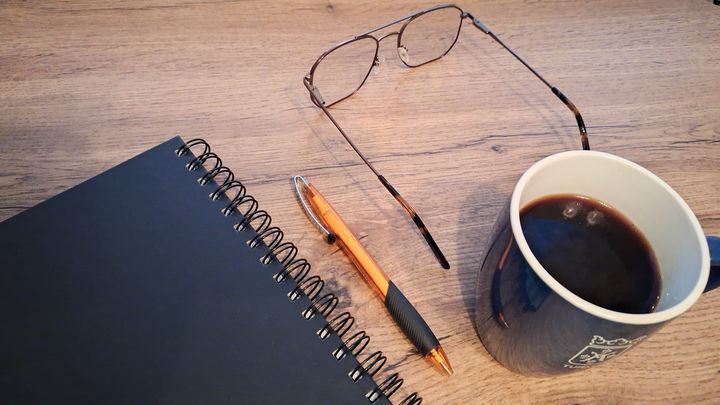 Kuvassa on opiskeluvälineitä aseteltuna pöydälle. Musta muistiinpanovihko, oranssi kynä, silmälasit ja kahvimuki, jossa on Turun kaupungin vaakuna